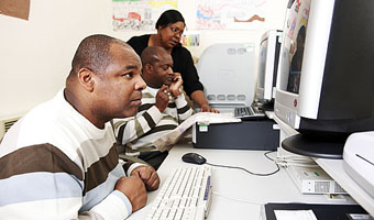 2 men at computers with woman leaning over one in backgroun